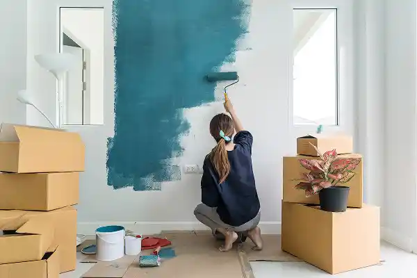 A person is painting a white wall teal with a roller in a room filled with moving boxes. A potted plant is on one box, conveying a sense of home renovation.