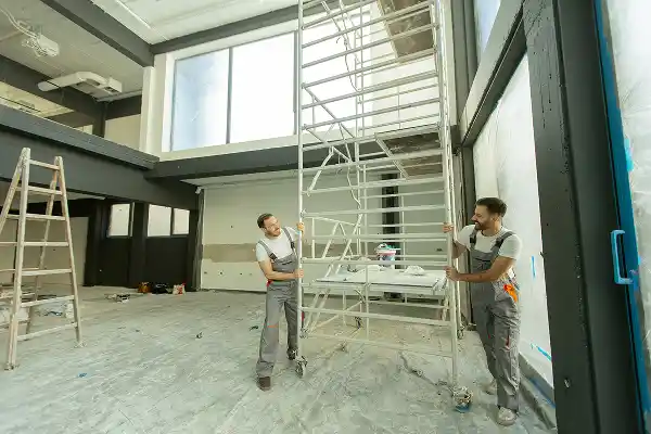 Two workers in overalls assemble a tall scaffolding inside a spacious, bright room. The setting conveys a mood of focused teamwork and construction.